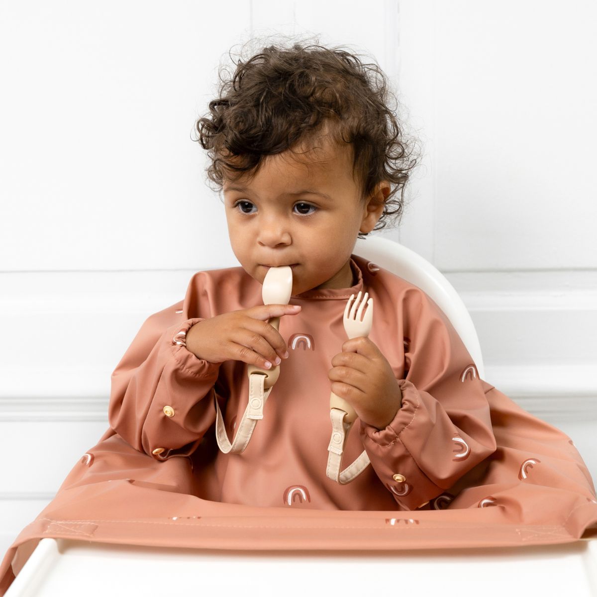 A baby feeding themselves with Bibado cutlery,
          wearing a Coverall attached to the highchair tray